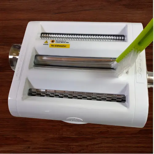 Cleaning a pasta roller with a soft brush to remove dried flour and dough buildup from the rollers and cutters without using water.