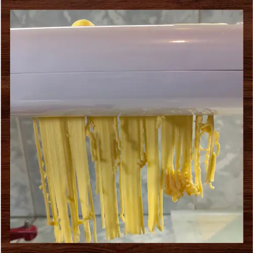 Fresh pasta strands emerging from a pasta cutter with torn, ragged noodles caused by dried pasta dough buildup inside the cutter.