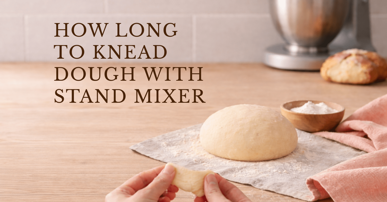 Hands stretching a small piece of dough for the windowpane test on a wooden counter, with a smooth dough ball on a floured cloth, a small bowl of flour, an apricot towel, and a stand mixer blurred in the background.