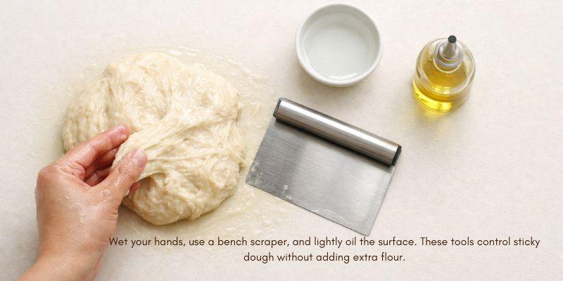 Sticky bread dough being lifted with wet fingertips on a lightly oiled countertop, with a bench scraper, water dish, and small oil bottle beside it (no flour used).